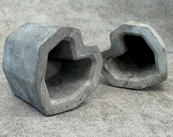 May include: Two gray, geometric planters with heart-shaped openings. The planters have a rough, textured surface and a modern, minimalist design. The planters are made of concrete and are suitable for indoor or outdoor use.