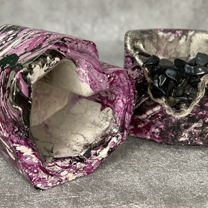 Könnte beinhalten: Zwei Beton-Pflanzer mit einem marmorierten Design in Lila, Schwarz und Weiß. Ein Pflanzer hat schwarze Steine ​​im Inneren.