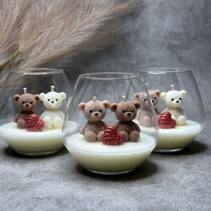 May include: Three glass candle holders each containing a white and brown teddy bear couple. The bears are holding a red heart. The candle holders are on a grey surface.
