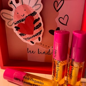 May include: Three pink and yellow tubes of 100% Lanolin Oil Lip Gloss by Dash Organics. The tubes are on a red wooden surface with a pink and white bee illustration.