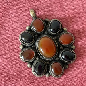 Op de afbeelding: Een ronde metalen hanger met een grote centrale ovale oranje steen, omringd door acht kleinere afwisselend zwarte en oranje ovale stenen. De metalen zetting heeft een getextureerde, donkere afwerking met kleine bolvormige accenten.