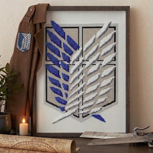 Puede incluir: Un bordado en punto de cruz enmarcado que presenta el emblema de las Alas de la Libertad con alas azules y blancas sobre un fondo beige. El diseño se muestra junto a una chaqueta marrón con un parche a juego y un componente de arma de metal.
