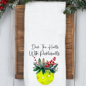 May include: White waffle-weave kitchen towel with the text "Deck The Halls With Pickleballs" and a watercolor pickleball design. The towel is displayed on a wooden holder, with evergreen branches and red berries in the background.
