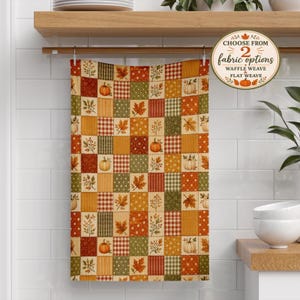 May include: A kitchen towel featuring a patchwork pattern of autumn-themed squares in shades of orange, green, yellow, and brown. The design includes illustrations of pumpkins, maple leaves, and plaid textures. A circular badge reads CHOOSE FROM 2 fabric options WAFFLE WEAVE & FLAT WEAVE.