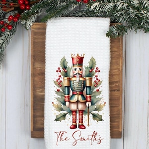 May include: White waffle-weave kitchen towel featuring a colorful illustration of a nutcracker soldier with a crown, holding a staff, and surrounded by holly and berries. The text "The Smiths" is written in a script font at the bottom.