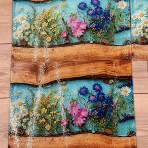 May include: Rectangular wooden serving boards with a light blue resin center, embedded with colorful dried flowers. The flowers include white daisies, yellow wildflowers, pink blossoms, and blue flowers. The wood has a natural, rustic appearance.