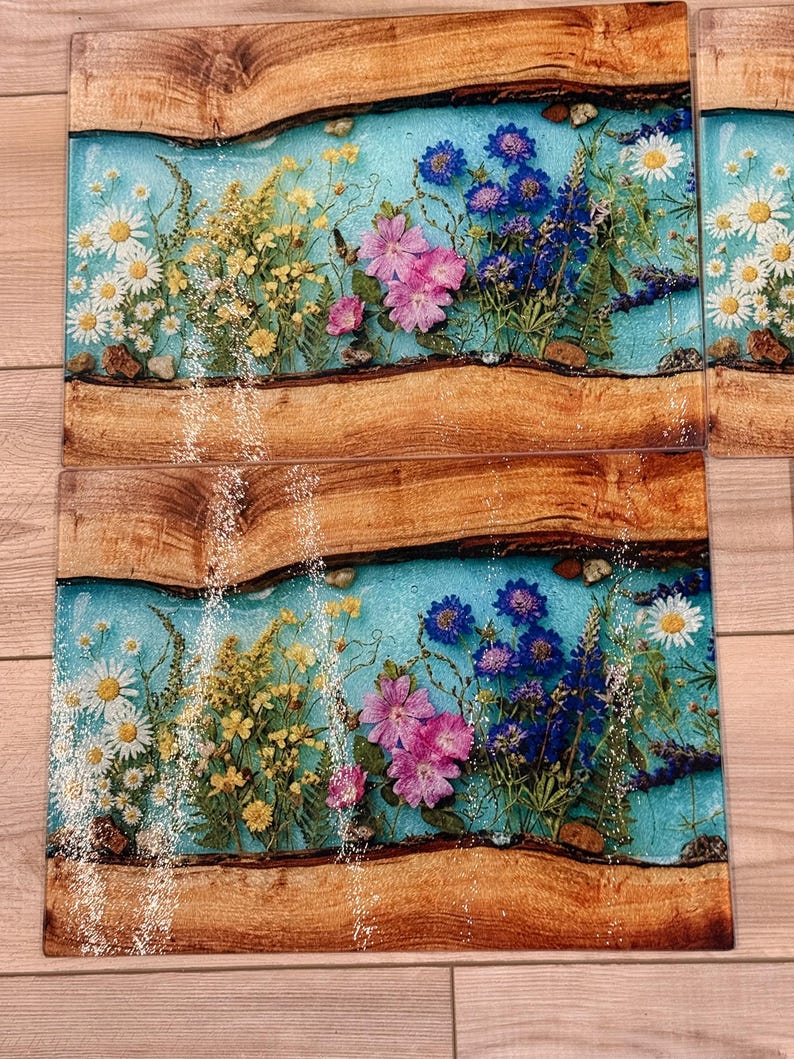 May include: Rectangular wooden serving boards with a light blue resin center, embedded with colorful dried flowers. The flowers include white daisies, yellow wildflowers, pink blossoms, and blue flowers. The wood has a natural, rustic appearance.