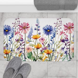 Könnte beinhalten: Eine rechteckige Badematte mit einem floralen Aquarelldesign. Die Matte zeigt eine lebendige Anordnung von Blumen in Rosa-, Blau-, Gelb- und Lilatönen vor einem weißen Hintergrund. Graue Hausschuhe sind im Vordergrund.