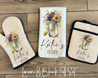 May include: A kitchen set consisting of an oven mitt, a waffle-weave towel, and a pot holder. Each piece is off-white with a black trim and features a graphic of a mason jar filled with wildflowers and the text Katie's KITCHEN.