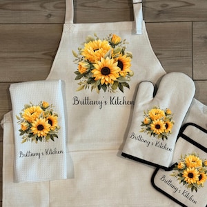 May include: A kitchen set featuring a white apron, oven mitt, pot holders, and a towel. Each item is decorated with a vibrant sunflower bouquet and the text "Brittany's Kitchen". The set is available to purchase individually or as a set.