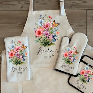 May include: A kitchen set featuring an apron, oven mitt, pot holder, and dish towel. Each item is beige and adorned with a floral design, butterflies, and the text "Bailey's Kitchen". The text "Available as a set or individually" is at the top.