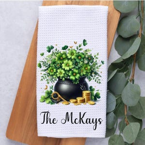 May include: White kitchen towel with a black pot filled with green shamrocks and gold coins. The text "The McKays" is printed below the pot.