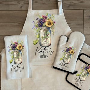 May include: A kitchen set featuring an apron, oven mitt, pot holder, and dish towel. Each item displays a floral design in a mason jar with the text "Katie's Kitchen". The text "Available as a set or individually" is at the top.