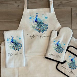 May include: A kitchen set featuring a peacock design. The set includes an apron, oven mitt, pot holder, and a kitchen towel. Each item has a cream-colored background with a blue and gold peacock illustration and the text "Taylor's Kitchen".