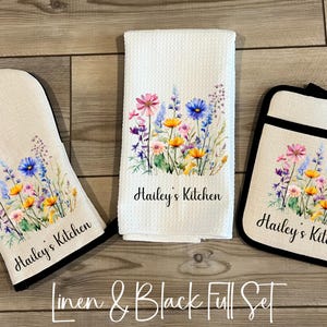 May include: A kitchen set featuring a white oven mitt, a white kitchen towel, and a white pot holder. Each item is decorated with a floral design in pink, blue, and yellow, with the text "Hailey's Kitchen". The set has black trim.