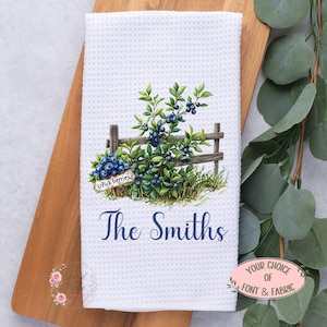 May include: A white waffle-weave kitchen towel featuring a watercolor illustration of a blueberry bush behind a wooden fence with a sign reading U-Pick berries and the dark blue text The Smiths printed in a cursive script font below the graphic.