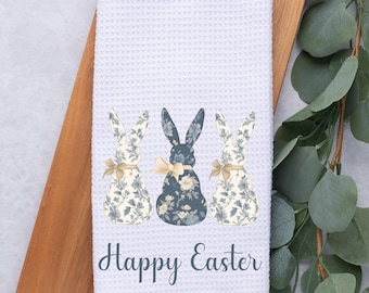 May include: White waffle-weave kitchen towel featuring three bunny silhouettes in blue floral patterns and tan bows. The central bunny is navy blue. The words "Happy Easter" are printed in a script font.