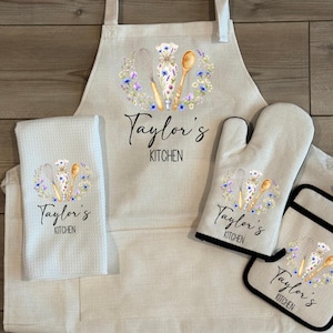 May include: A beige kitchen set featuring an apron, oven mitt, pot holder, and a towel. Each item is decorated with a floral wreath design and the text "Taylor's Kitchen". The apron has adjustable straps.
