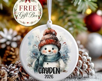May include: A round white ceramic ornament featuring a watercolor illustration of a snowman wearing a brown knit hat and a blue scarf. The name CAYDEN and the year 2026 are printed in black text below the snowman. A white ribbon is attached.