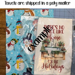 Pu&ograve; includere: Un asciugamano da cucina bianco con testo rosso che recita "There's No Place Like Home For The Holidays". L'asciugamano &egrave; avvolto in un sacchetto di plastica trasparente e posizionato su carta da regalo blu con un motivo a pupazzo di neve.