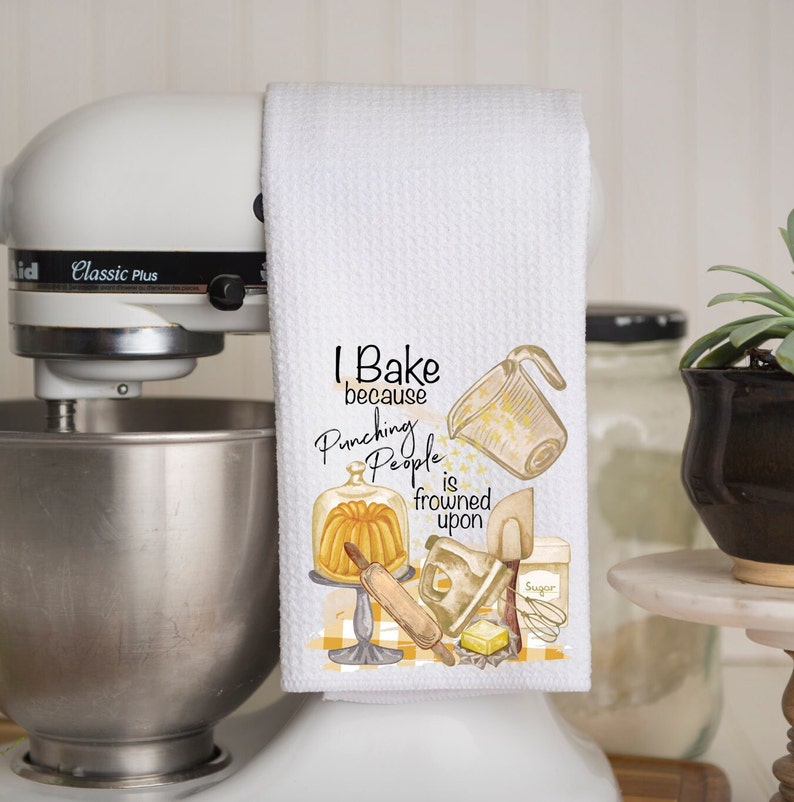 Pu&ograve; includere: Asciugamano da cucina bianco con un'illustrazione gialla e marrone di utensili da cucina e il testo "I Bake because Punching People is frowned upon".