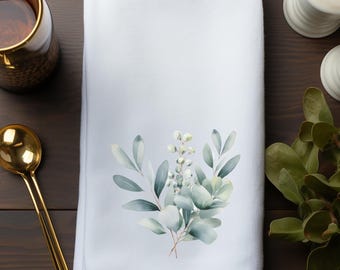 May include: A white rectangular kitchen towel featuring a central watercolor illustration of green and blue-toned eucalyptus leaves and small white berries. The fabric is smooth and folded, displaying a clean, crisp appearance with botanical art.