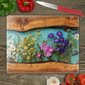 May include: A rectangular wooden cutting board with a turquoise resin center, embedded with colorful dried flowers. A red-handled knife rests above the board. Green and red bell peppers and tomatoes are also visible.