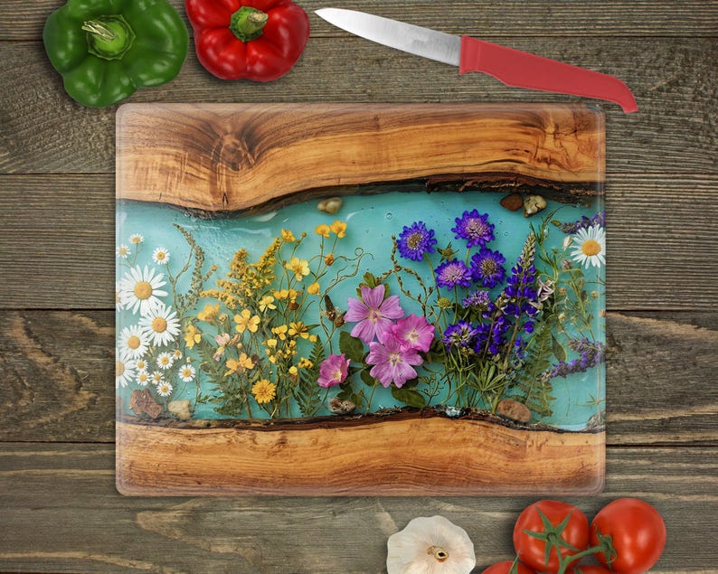 May include: A rectangular wooden cutting board with a turquoise resin center, embedded with colorful dried flowers. A red-handled knife rests above the board. Green and red bell peppers and tomatoes are also visible.