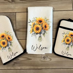 May include: A set of three kitchen items featuring a sunflower bouquet design. The set includes a potholder, an oven mitt, and a tea towel. Each item has the name "Wilson" printed on it.