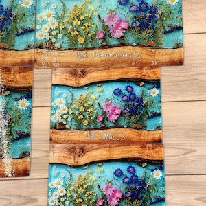 May include: Rectangular cutting boards with a blue resin river design, featuring pressed flowers in white, yellow, and pink. The boards have a wooden border and the text "The Plamondon's" and "THE SMALL'S".