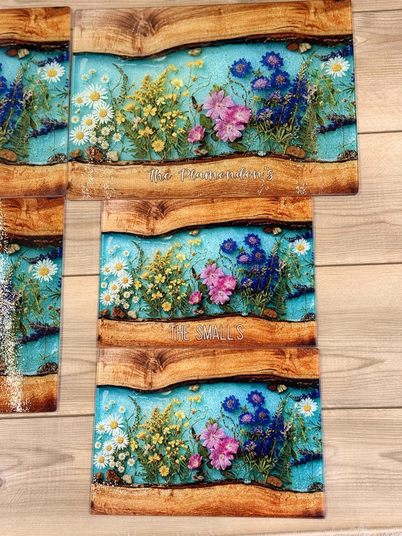 May include: Rectangular cutting boards with a blue resin river design, featuring pressed flowers in white, yellow, and pink. The boards have a wooden border and the text "The Plamondon's" and "THE SMALL'S".