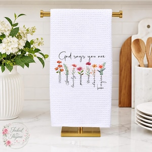 May include: A white waffle-weave kitchen towel hanging on a gold metal rack. The towel features a floral design with the text "God says you are" followed by the words "Unique, Special, Lovely, Precious, Strong, Chosen, Forgiven" in black script.