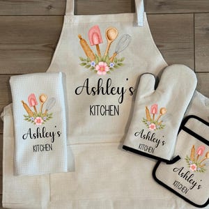 May include: A kitchen set featuring an apron, oven mitt, pot holder, and a kitchen towel. Each item is off-white and decorated with a watercolor design of kitchen utensils and the text "Ashley's Kitchen".