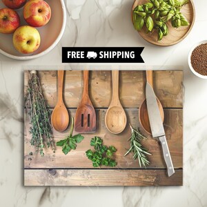 May include: A rectangular glass cutting board featuring a printed image of three wooden kitchen utensils, a silver chef's knife, and various fresh green herbs arranged on a rustic wooden plank surface. The text FREE SHIPPING is printed in white.