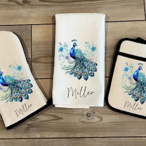 May include: A set of three kitchen items featuring a peacock design. The set includes a tea towel, a pot holder, and an oven mitt. Each item is made of a natural fabric and features a blue and green peacock with a gold and white floral design. The items are personalized with the name "Miller".