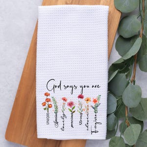May include: White kitchen towel with a floral design and the text "God says you are" with the words "unique, special, lovely, precious, strong, chosen, forgiven" written vertically below the flowers.