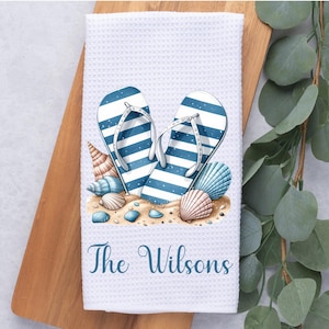 May include: White kitchen towel with a blue and white striped flip-flop design and seashells. The text "The Wilsons" is printed on the towel.