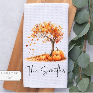 May include: White waffle-weave kitchen towel featuring a watercolor illustration of a tree with orange and yellow leaves, pumpkins, and the text "The Smiths" in black script. The towel has a fall or autumn theme.