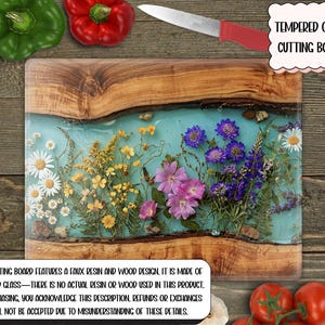 May include: Tempered glass cutting board with a faux resin and wood design. The board features a light blue resin center with embedded dried flowers. A knife with a red handle rests on the board. The text on the board reads: "TEMPERED GLASS CUTTING BOARD."