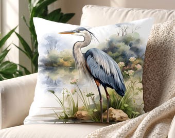 May include: A square throw pillow featuring a watercolor illustration of a blue heron standing near a calm lake. The bird has grey and blue feathers with orange accents on its legs. The scene includes green reeds, small pink flowers, and rocks.