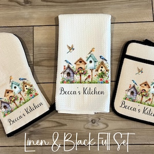 May include: A kitchen set featuring a white oven mitt, a white hand towel, and a black-trimmed pot holder. Each item displays a colorful birdhouse design with birds and flowers, and the text "Becca's Kitchen". The set is on a wooden surface.