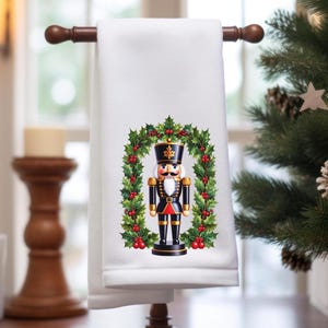 May include: White hand towel with a festive design featuring a nutcracker soldier within a holly wreath. The nutcracker is dressed in a black uniform with gold accents and a tall hat. Red berries and green leaves complete the holiday theme.