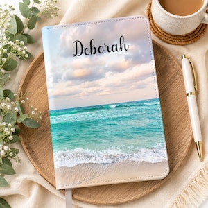 May include: A rectangular notebook featuring a printed image of a turquoise ocean with white waves crashing onto a sandy beach under a cloudy sky. The name Deborah is printed in black cursive script on the upper portion of the notebook cover.