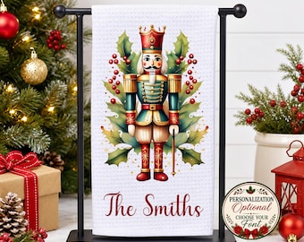 May include: A white waffle-weave kitchen towel hanging on a black metal stand, featuring a colorful nutcracker illustration surrounded by green holly leaves and red berries, with the custom text The Smiths printed in red script below.