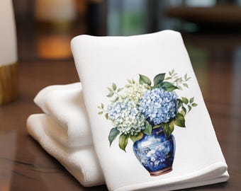 May include: A folded white hand towel featuring a printed graphic of a blue and white ceramic vase filled with light blue and white hydrangea flowers and green leaves. The towel has a soft, textured fabric surface and a clean, finished hem.