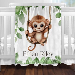 May include: White baby blanket featuring a smiling cartoon monkey swinging from a tree branch. The blanket has green leaf accents and the name "Ethan Riley" printed in black. The blanket is displayed on a white crib.