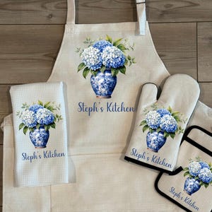 May include: A kitchen set featuring an apron, oven mitt, pot holder, and a towel. Each item displays a blue and white floral design with the text "Steph's Kitchen". The set is made of a light-colored fabric.