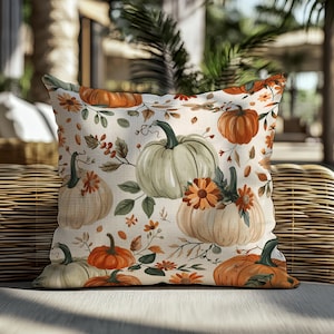 May include: A square decorative pillow with a cream background featuring a repeating pattern of pumpkins in shades of orange, green, and cream, along with orange flowers and green leaves. The pillow is placed on a wicker chair.