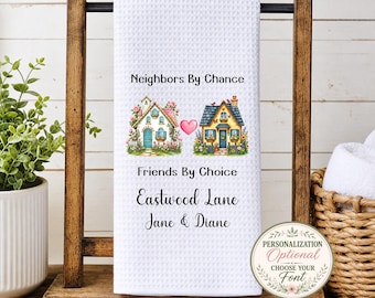 May include: A white waffle-weave kitchen towel featuring two illustrated houses separated by a pink heart. Text reads: Neighbors By Chance, Friends By Choice, Eastwood Lane, and Jane & Diane. The towel hangs on a wooden stand.