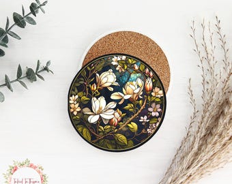 May include: A round coaster featuring a floral design with white magnolia blossoms, green leaves, and small pink flowers against a dark blue background. The coaster is placed on a white ceramic base with a cork backing. Decorative sprigs and foliage surround the coaster.